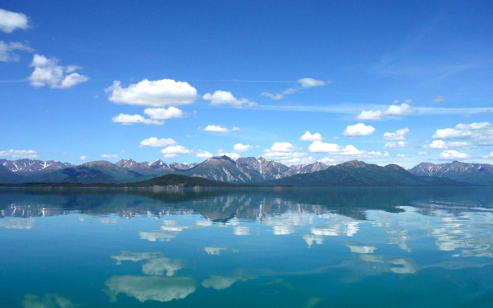 Photo of blue sky with fluffy white clouds reflect in calm lake with mountains in the background.